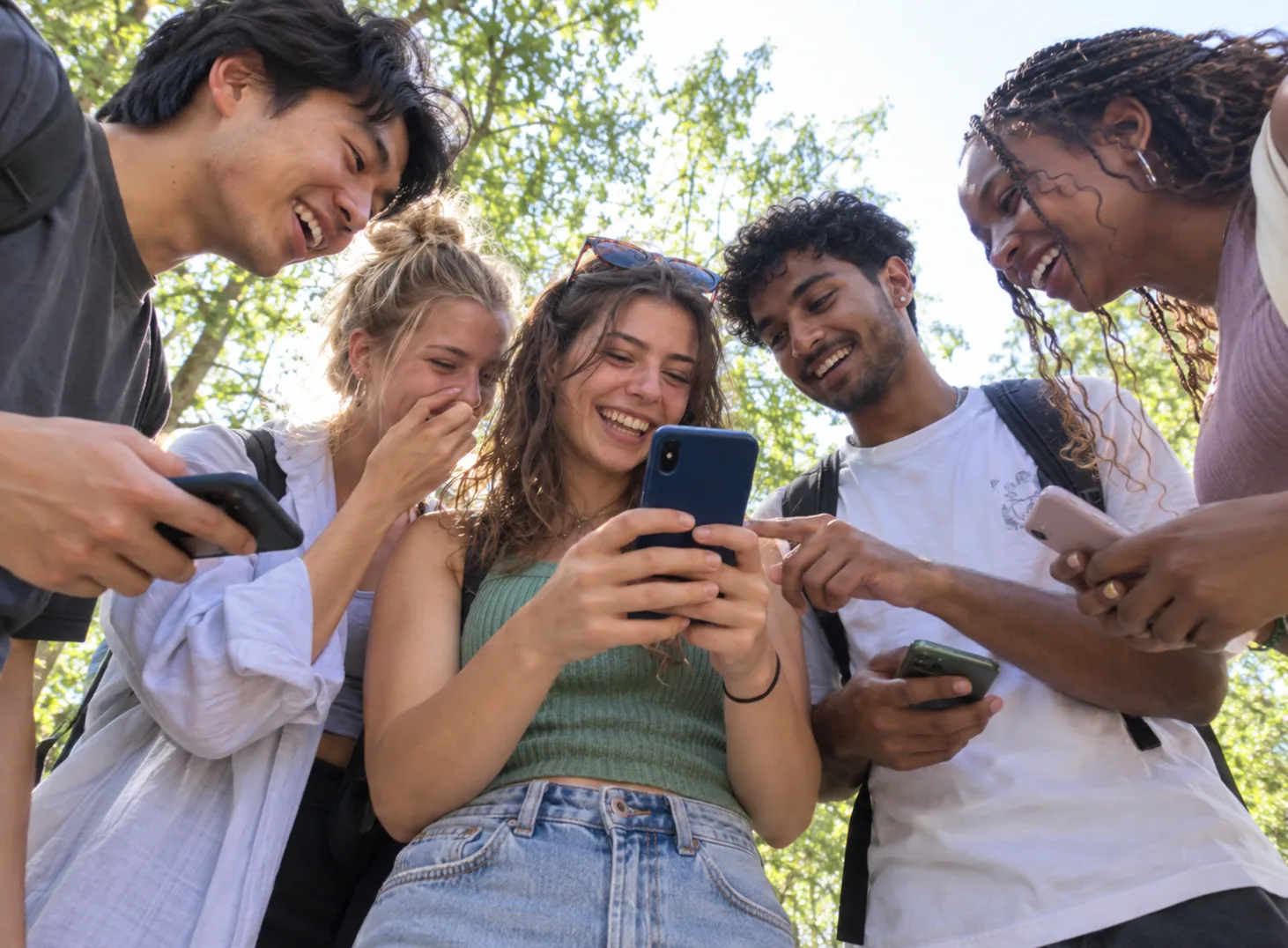 Des étudiants rient en regardant un téléphone, dehors sous les arbres, pendant un jeu de piste