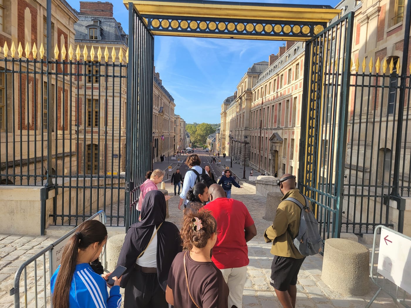 Des visiteurs franchissent les grilles dorées du château de Versailles et entrent dans la rue de l'Indépendance américaine